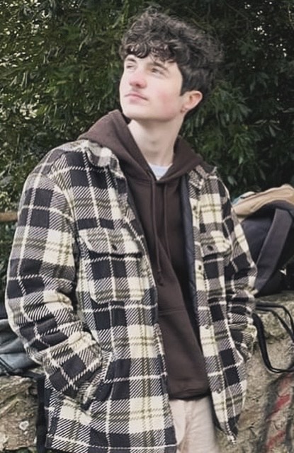 C'est un jeune homme au teint clair et aux cheveux bouclés. Il porte une veste à carreaux noirs et beiges sur un sweat à capuche marron, avec un pantalon clair. Ses mains sont dans les poches de cette veste, et un sac est posé sur un muret à côté de lui. Un arbre est en arrière-plan.